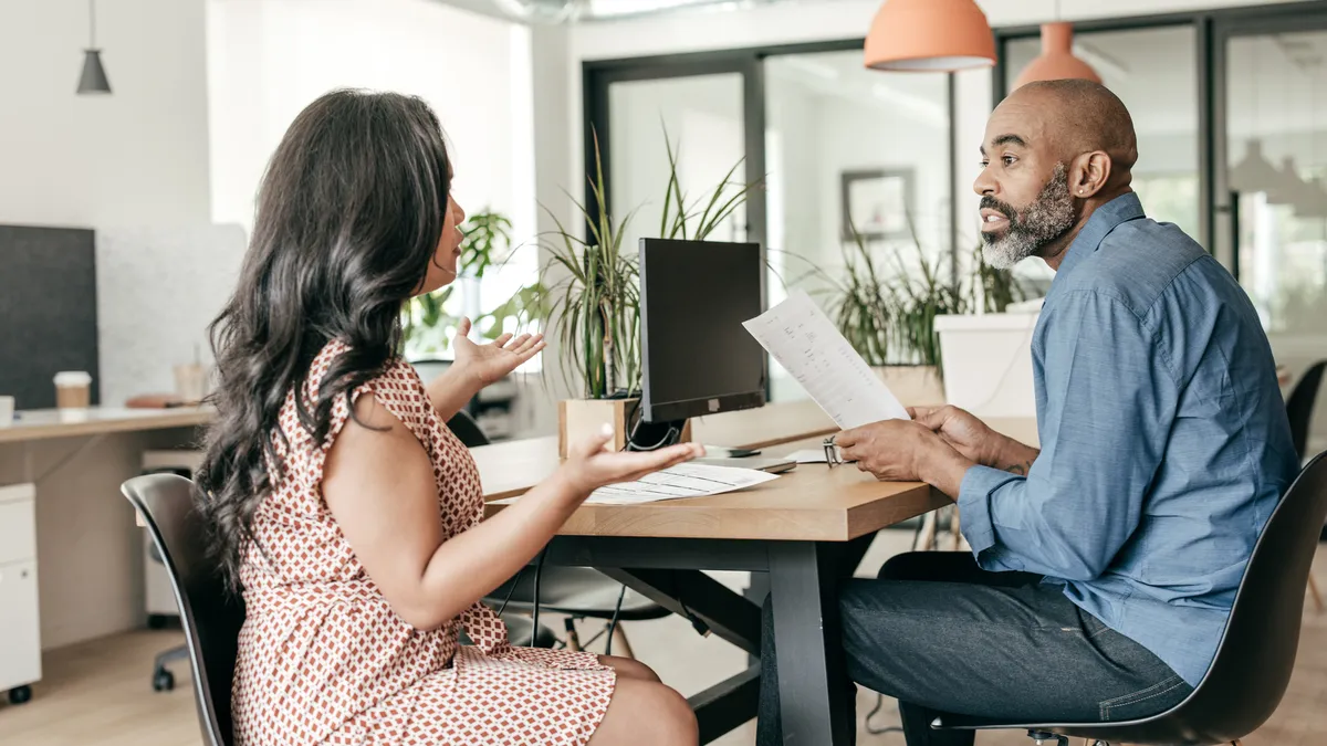 Two businesspeople sit across from each other, engaged and talking. One is holding paperwork