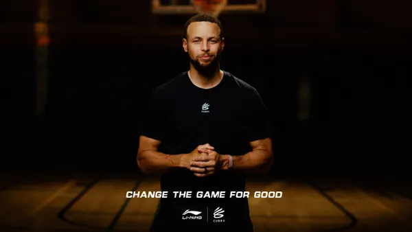 Steph Curry stands in a black T-shirt in front of a shadowed basketball court.