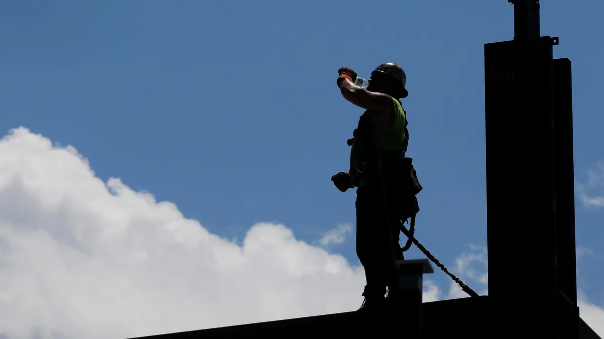 A construction worker atop a steel beam drinks from a bottle of water.