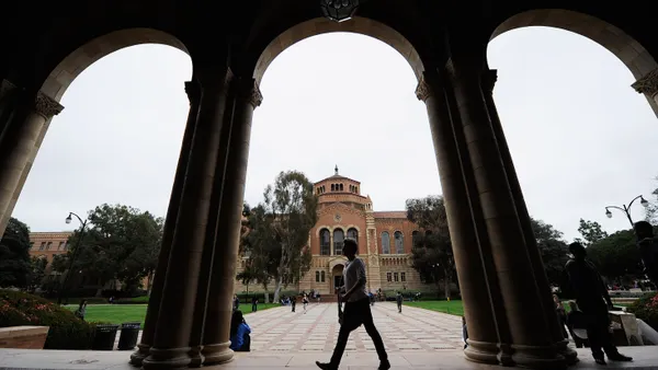 A student walks in front of arches, with a campus building in the background, on a college campus.