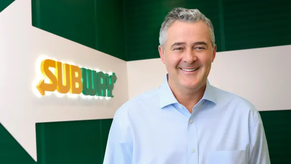 A smiling person stands in front of a green background with a yellow "Subway" logo visible.