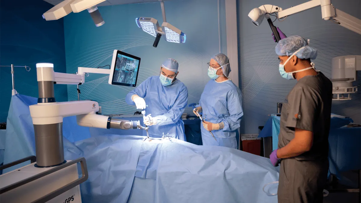 A person fits a tool in a robot arm while two other people wearing masks, caps and gloves stand around a hospital bed.