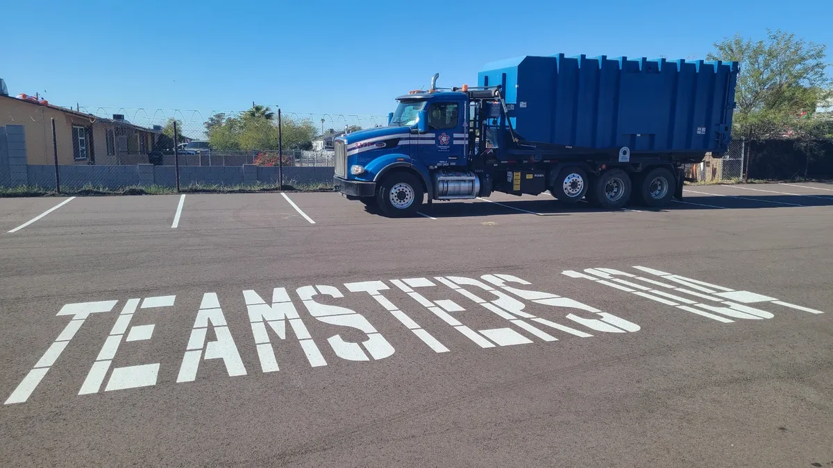 A waste truck sits in a parking lot with the words 'TEAMSTERS 104' painted on the pavement in front of it.