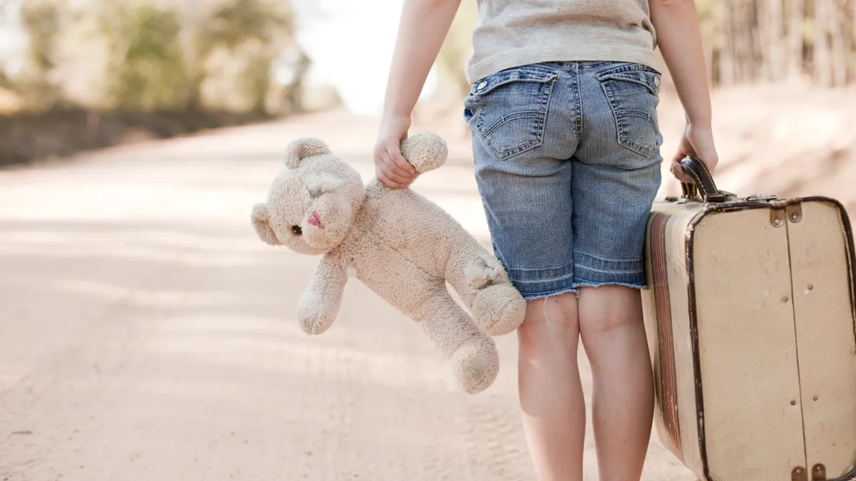 A child with back toward camera stands, shown below the shoulders, near a road with trees. In one hand is a worn-down teddy bear. In the other hand is a suitcase.