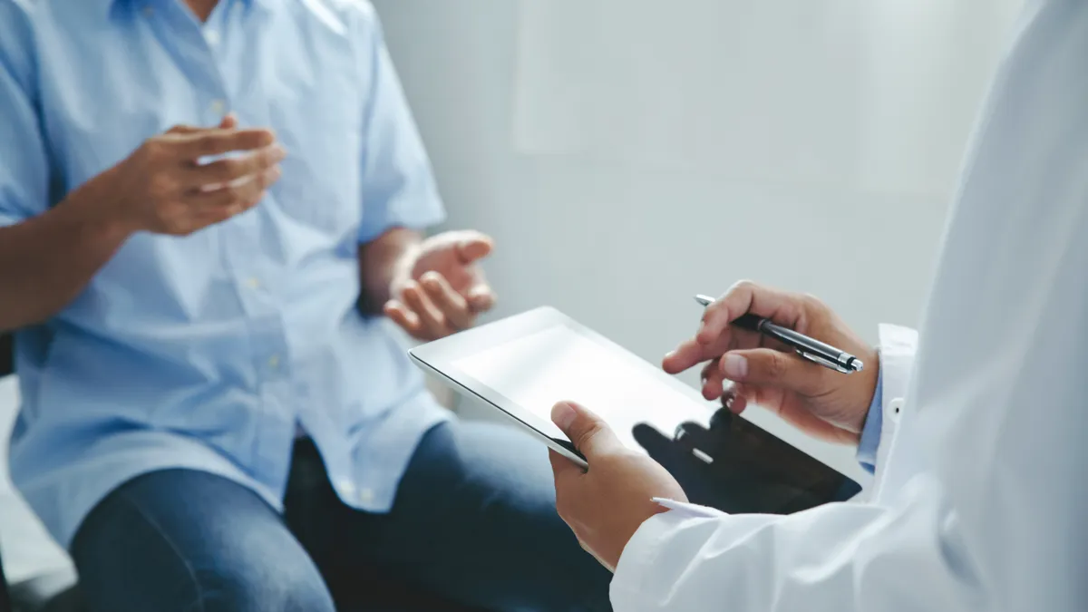 A person in a white lab coat holds an Ipad and pen and sits across from someone gesturing with their hands.