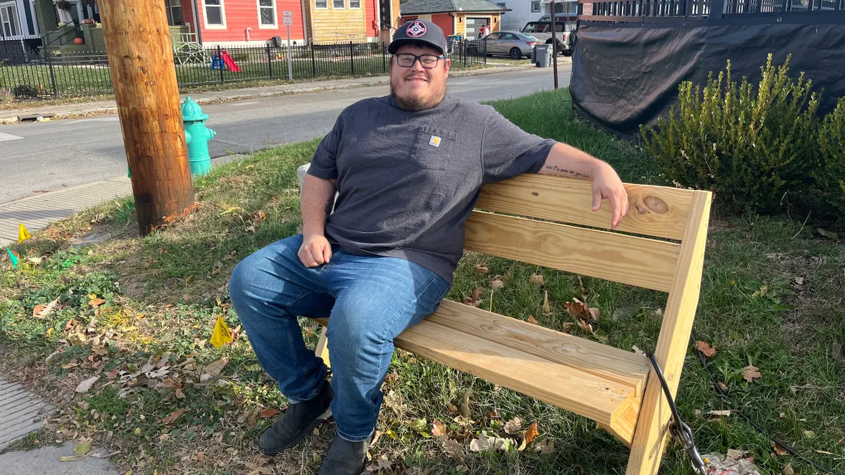 A person sits on a wooden bench on the grass with houses behind.