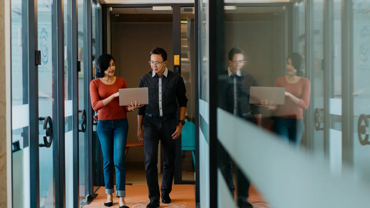 Two tech office workers walk down a hall looking at a laptop.