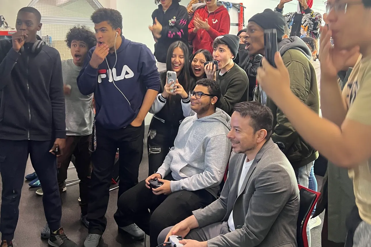 Students excitedly stand around a principal and student who are playing a game in a high school's video game club.