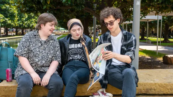 Three students sit at a bench flipping through a newspaper.