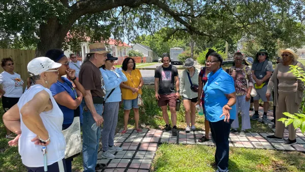 A person wearing a blue shirt and black pants talks to a group of people standing in a yard under a tree.