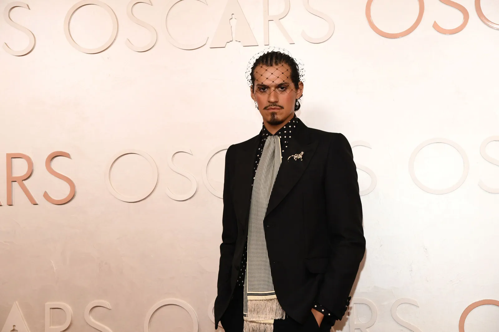 A man in a suit, ascot and birdcage veil stands on the red carpet.