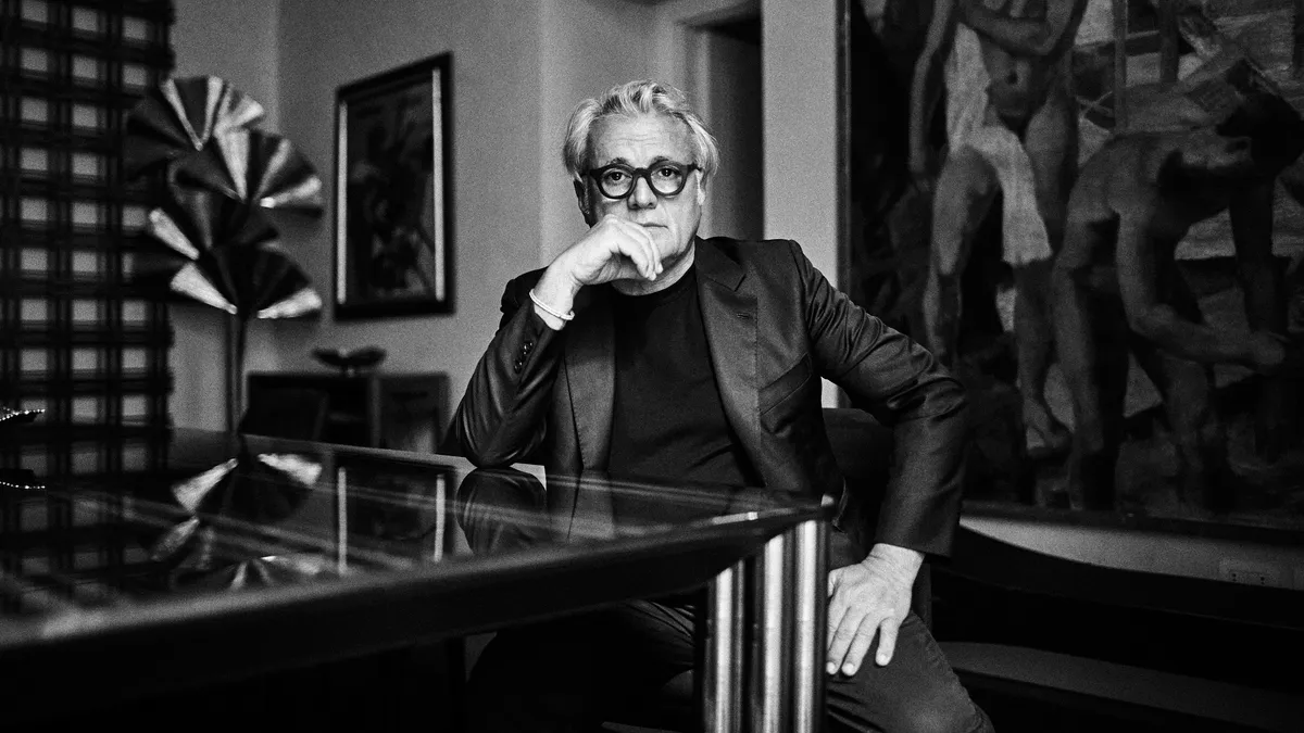 In a black-and-white photo, a man with white hair wears a black suit jacket and black T-shirt and leans on one hand while sitting at a glass table. A large painting is behind him.