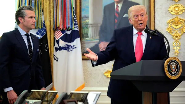 Two people in three-piece suits, with one standing behind a podium speaking and the other to the side looking on.