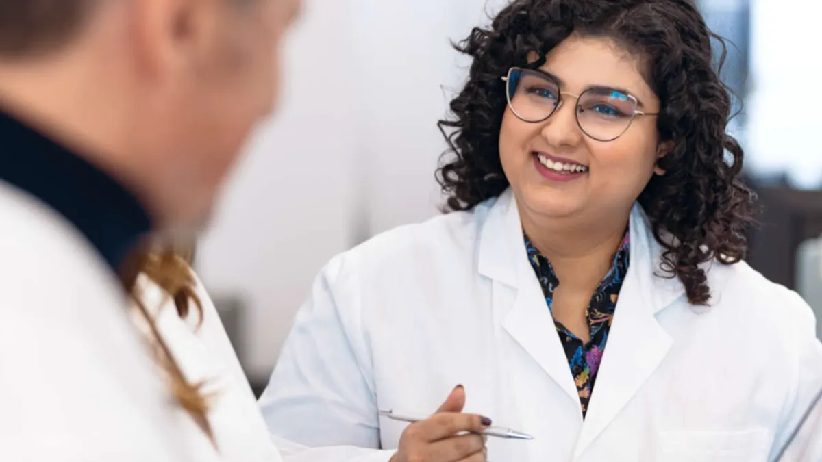 Smiling woman wearing glasses and a white lab coat engaging with other people.