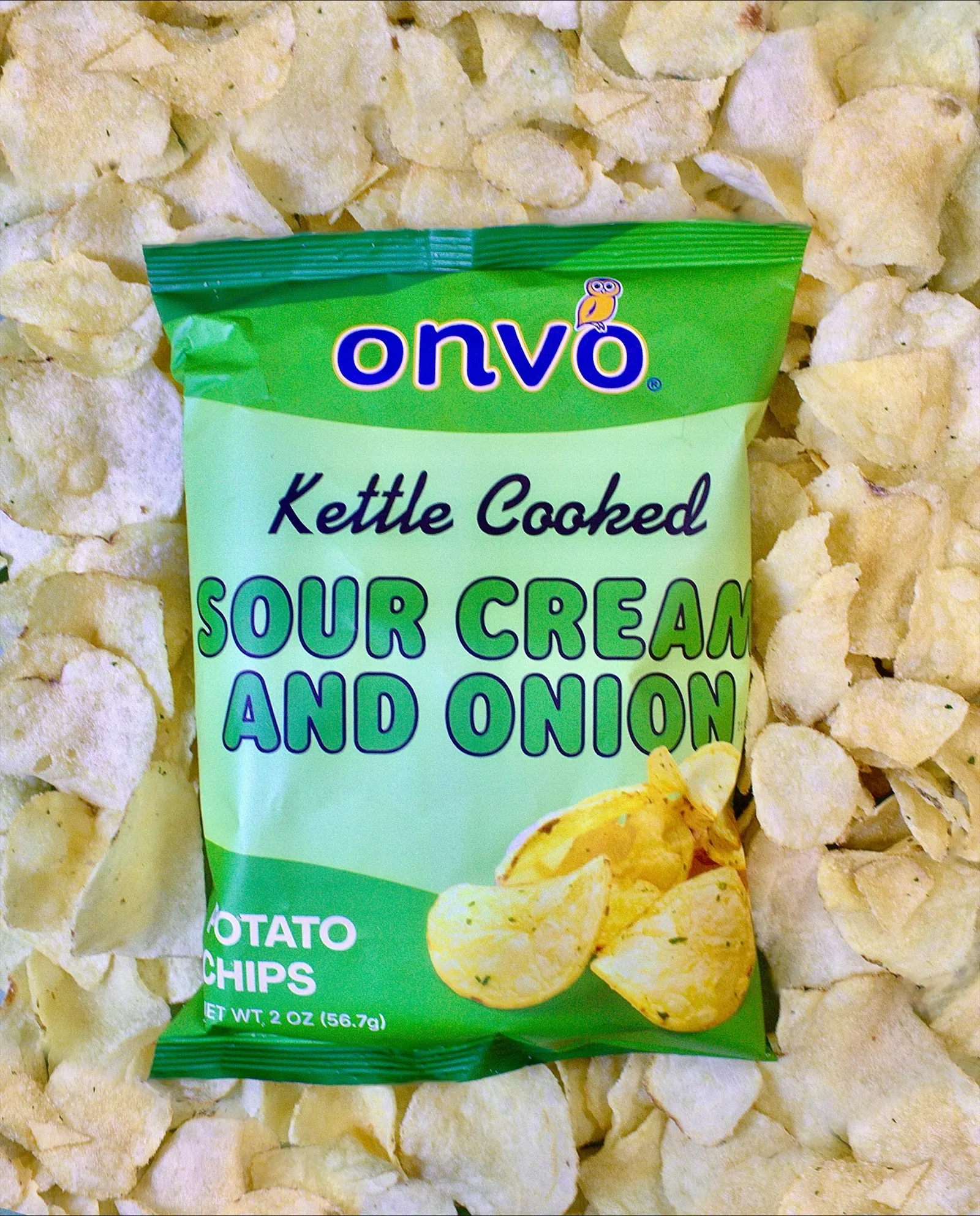 A bag of potato chips is lying atop a pile of potato chips. Text on the bag says Onvo kettle cooked sour cream and onion potato chips.