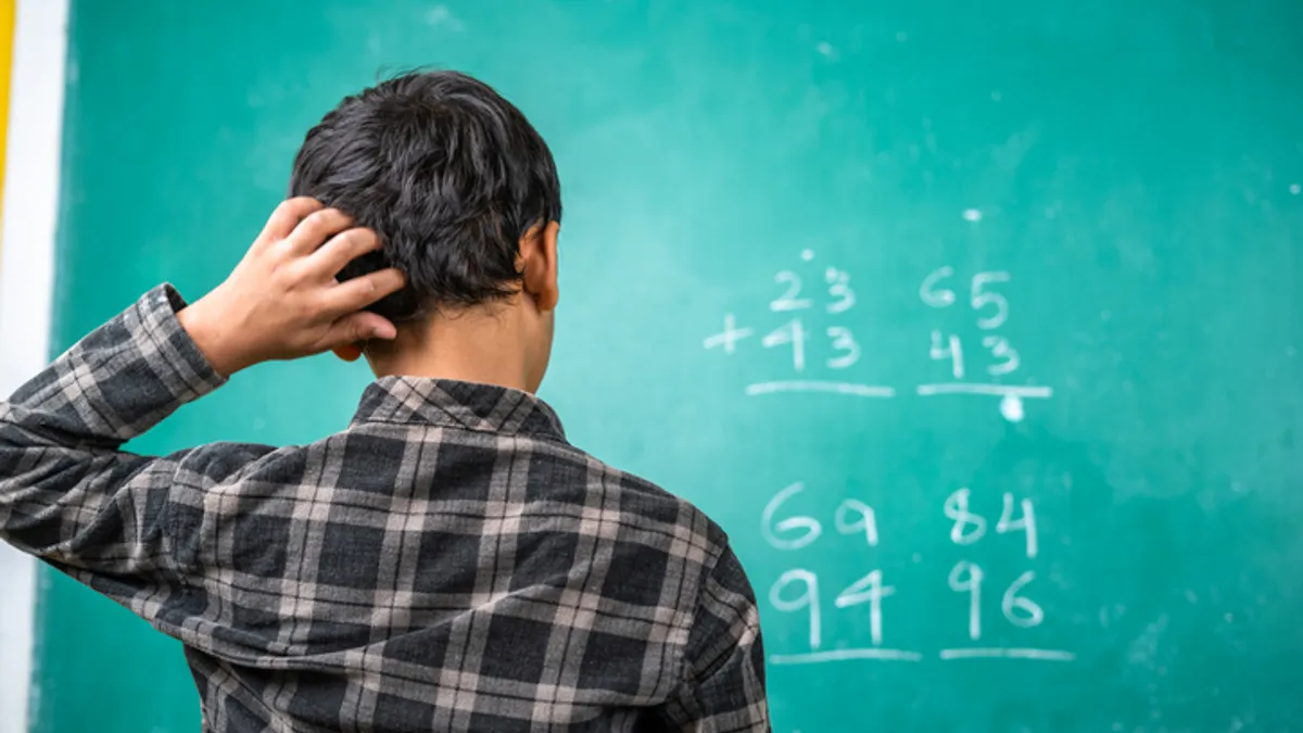 A student with their back to the camera faces a green chalk board. On the board are math problems. The student has their hand on their head.