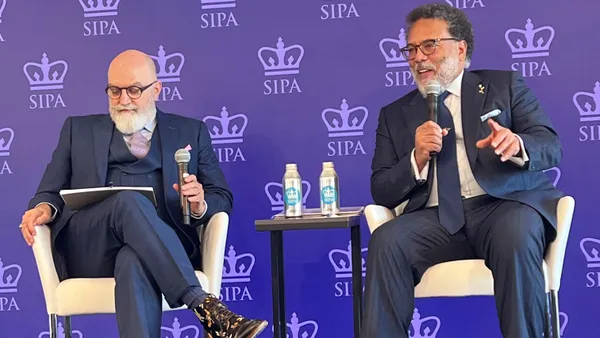 Two men in chairs on a stage hold microphones in front of a purple background with crowns and a SIPA logo