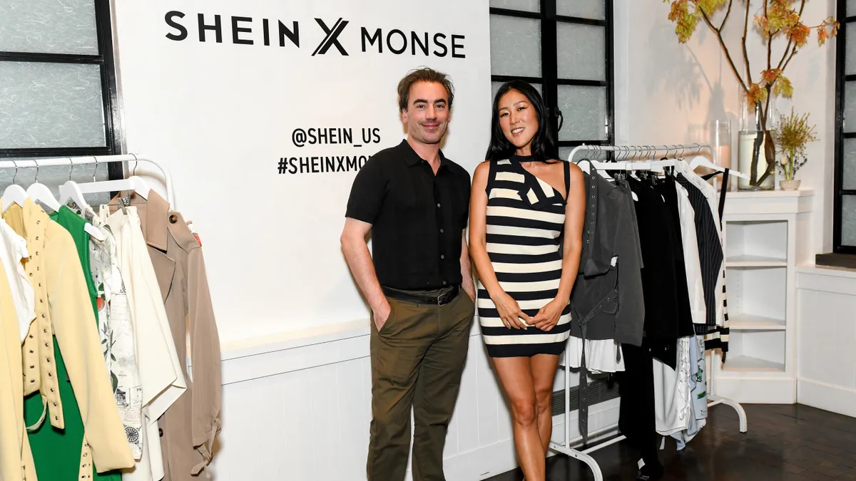 Two designers stand in front of their work, racks of clothing, against a wall which says SHEIN X MONSE