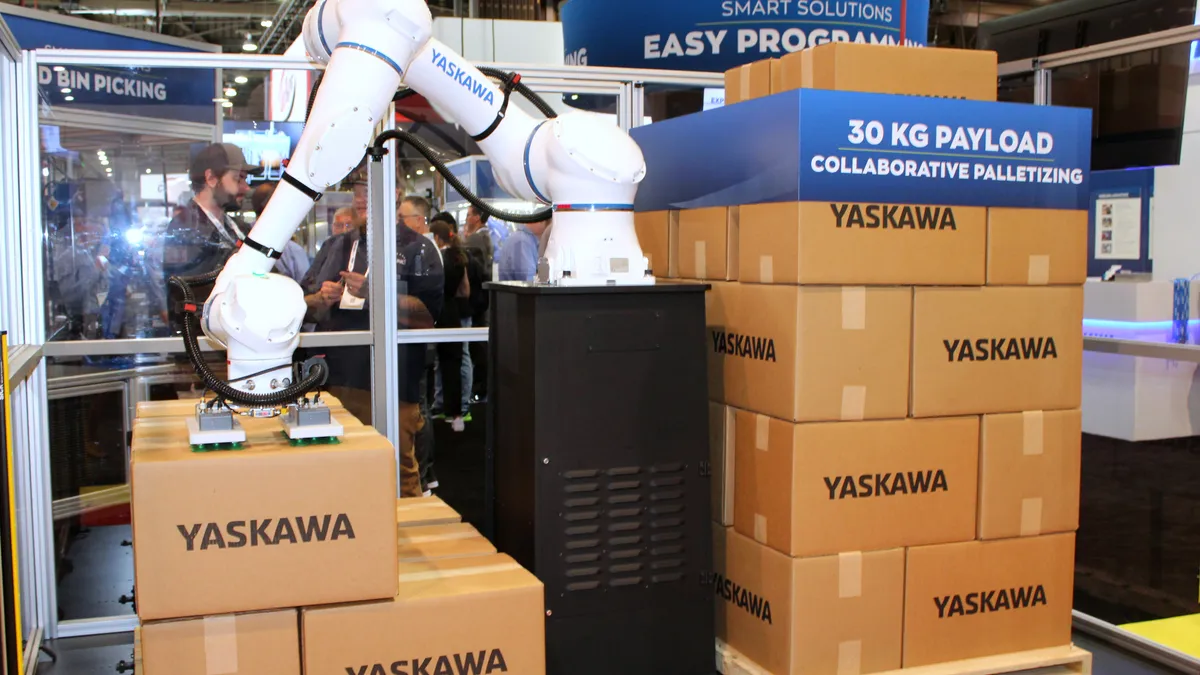 A white robot arm picks up cardboard boxes at a booth display at a conference.