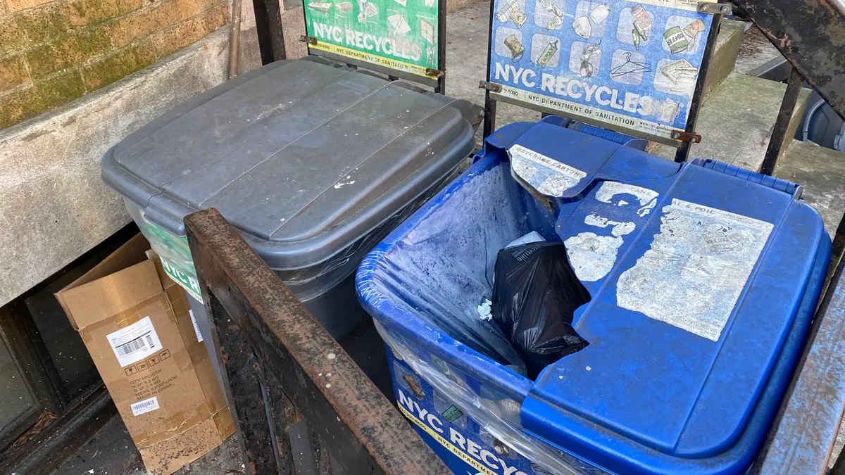 Brown and blue bins for waste and recycling are corralled in front of a building. The recycling bin's lid has been smashed in.