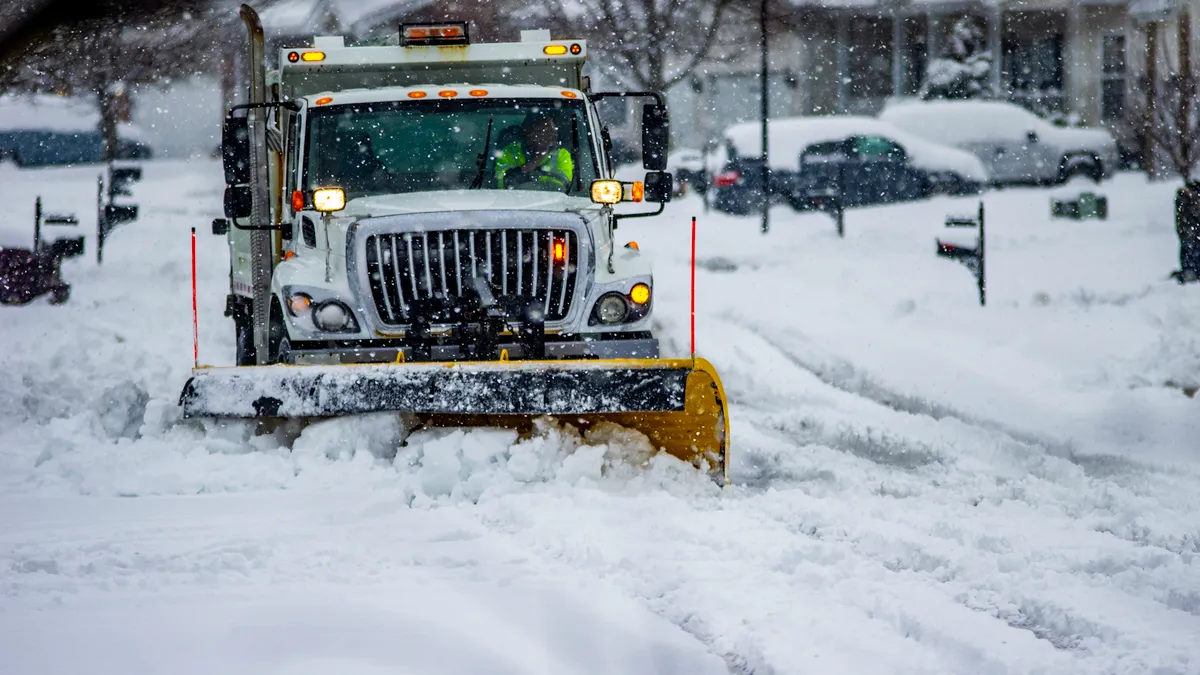 A snowplow on a large service truck pushes multiple inches of snow toward the viewer. The plow is on a suburban street while snow continues to fall.