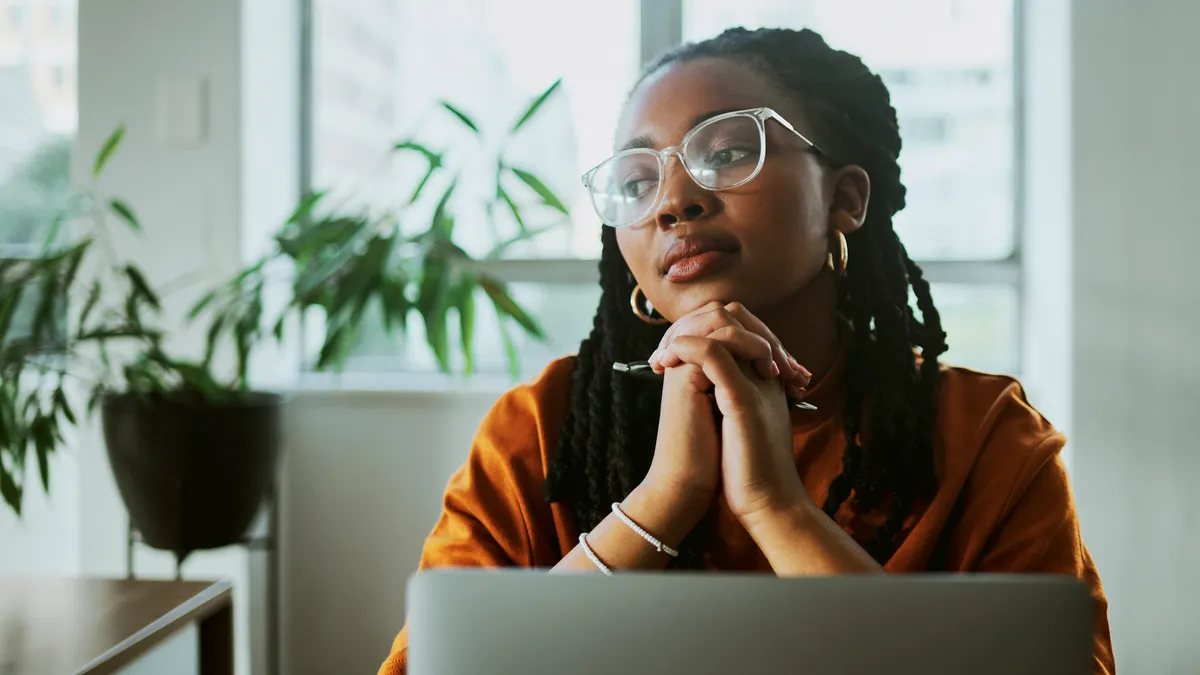 A Black feminine person with braids sits and thinks; they're deep in contemplation as they look off to the side over their laptop