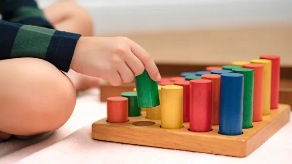 A close up of a child's hand and knee shows the hand placing a wooden column on a board with colorful wooden columns