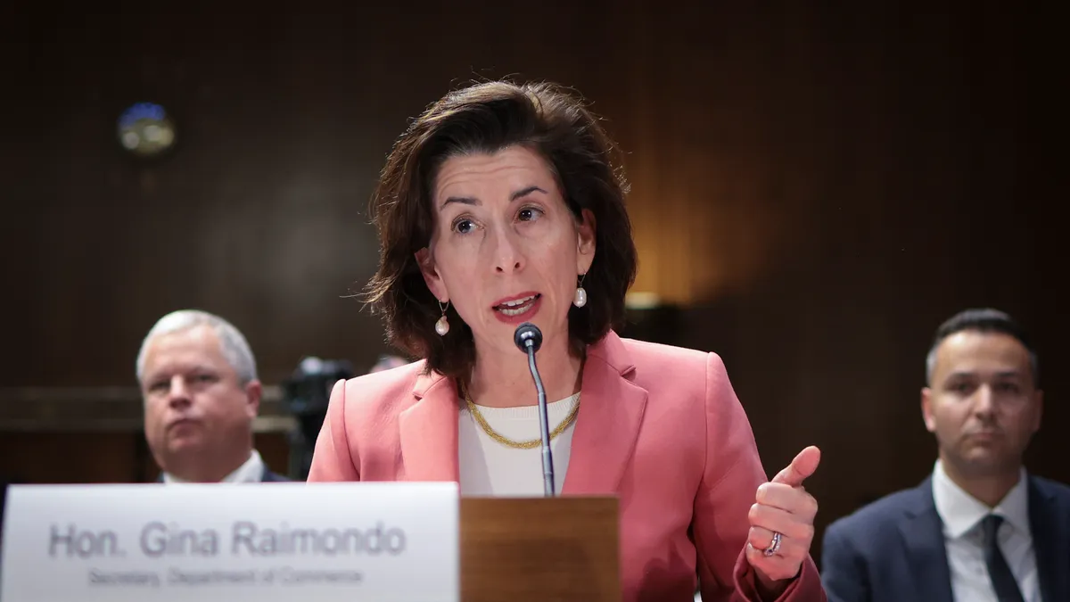 U.S. Secretary of Commerce Gina Raimondo in a peach blazer and white blouse speaking with a mic in front of her.