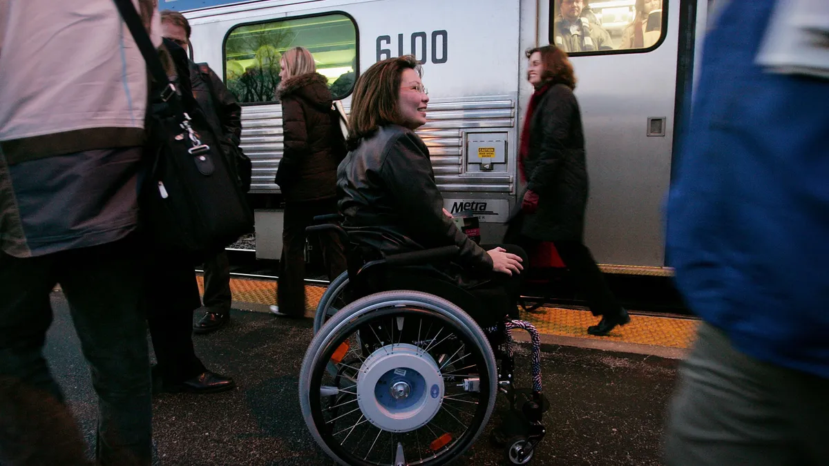 A woman in a wheelchair at a train station with several other people walking by, with a train behind them.