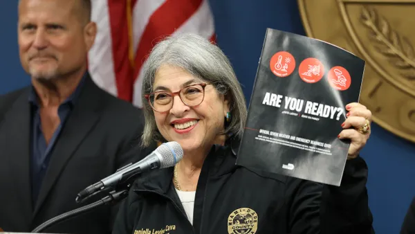 A person with gray hair wearing a jacket with a "Miami-Dade County" patch at a microphone holding up a brochure that says "ARE YOU READY?"