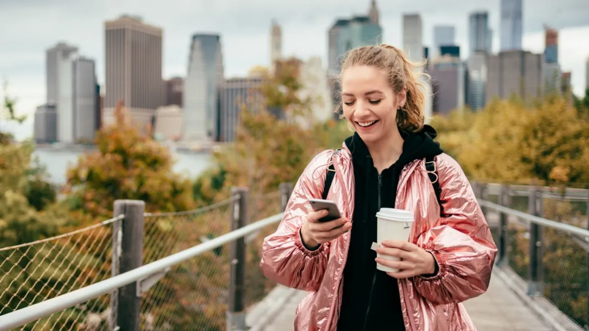 Person walking on bridge, texting on smartphone with one hand and holding a coffee with the other.