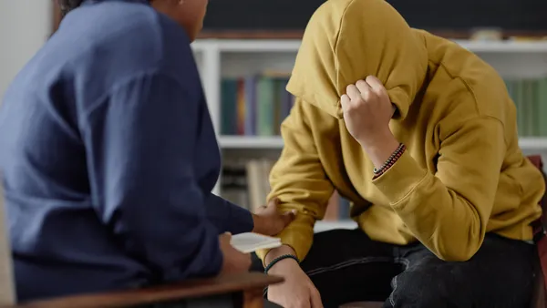 An adult, with their back toward the camera, is sitting in front of a student. The student is facing the adult and has a hood over their head hiding their face.
