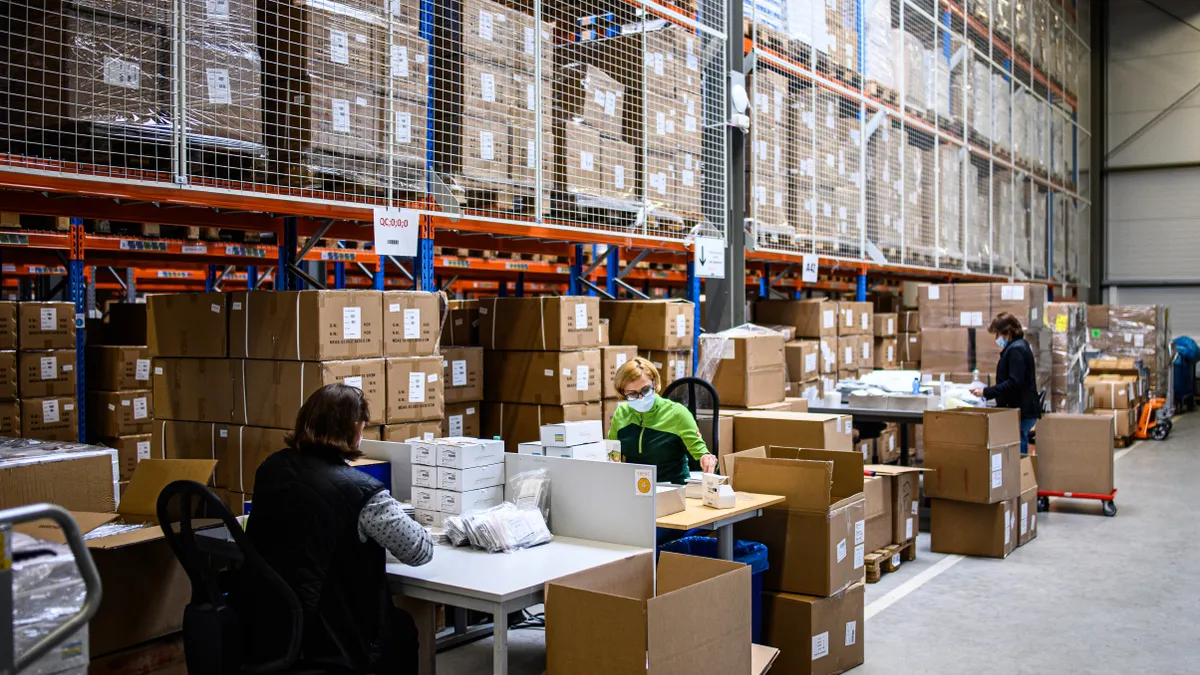 Workers sit a a manufacturing facility for rapid COVID-19 antigen tests. The workers are sitting at tables and surrounded by packages.