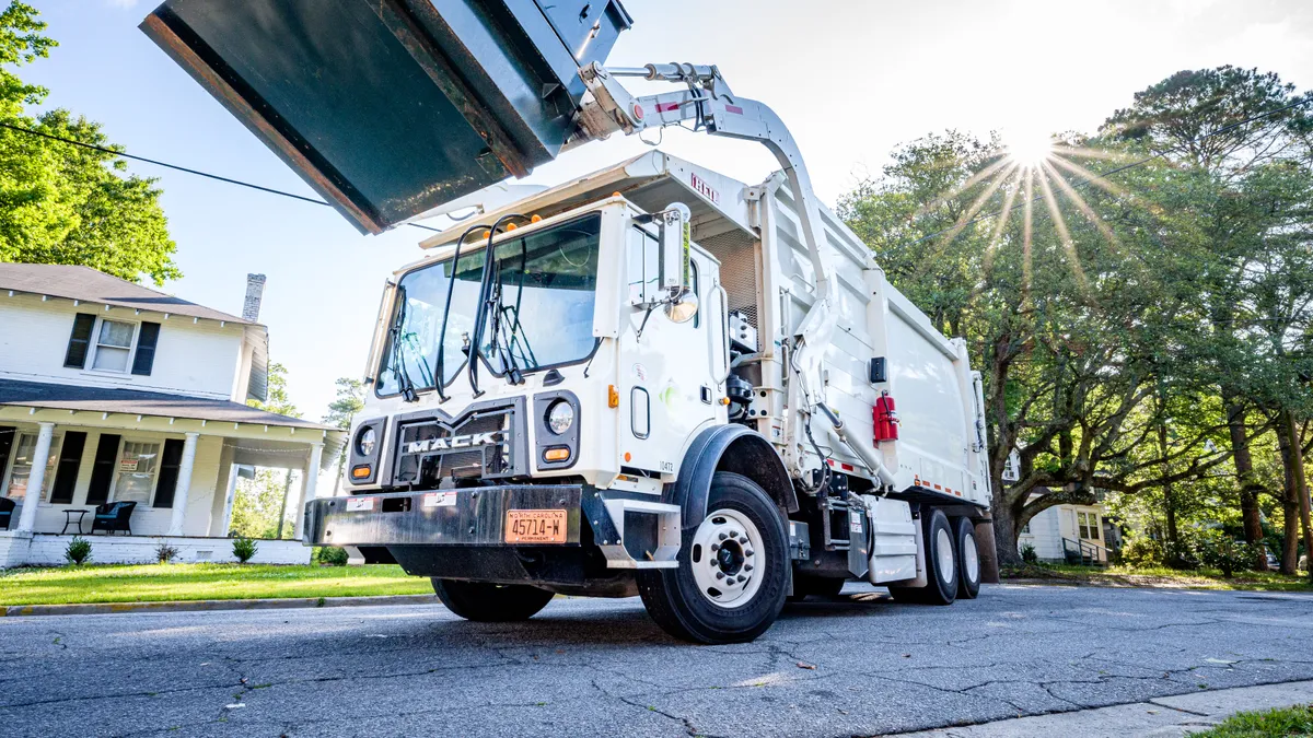 A front-loading garbage truck lifts a dumpster on a neighborhood street on a sunny day.