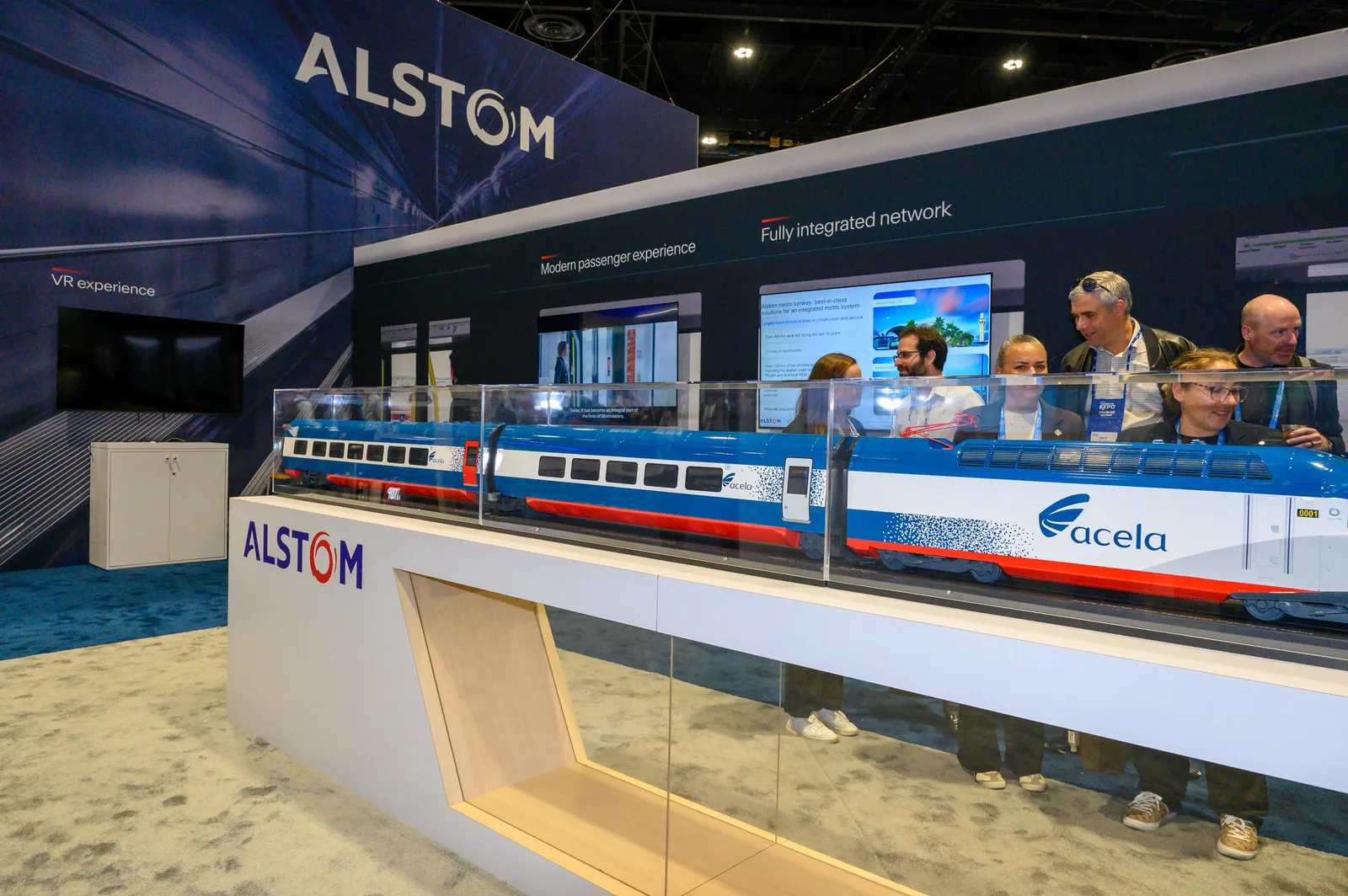 People look at a train model in a case with a sign reading "Alstom."