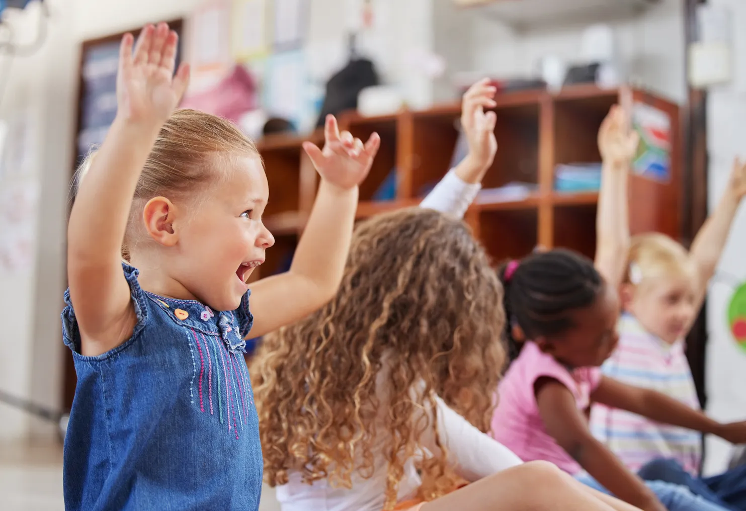 A handful of young children are seated on the floor in a row. They are looking forward and some have their arms raised.