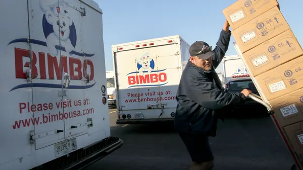 Bimbo Bakeries trucks are parked on the left, while a delivery man pulls a load of boxes to load.