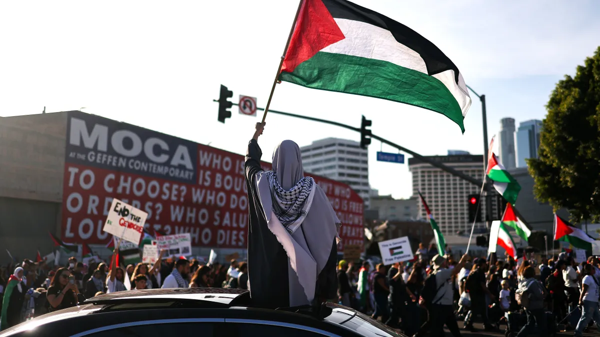 A picture of a person in a hijab, standing out of the sunroof of a car, waving Palestine's flag