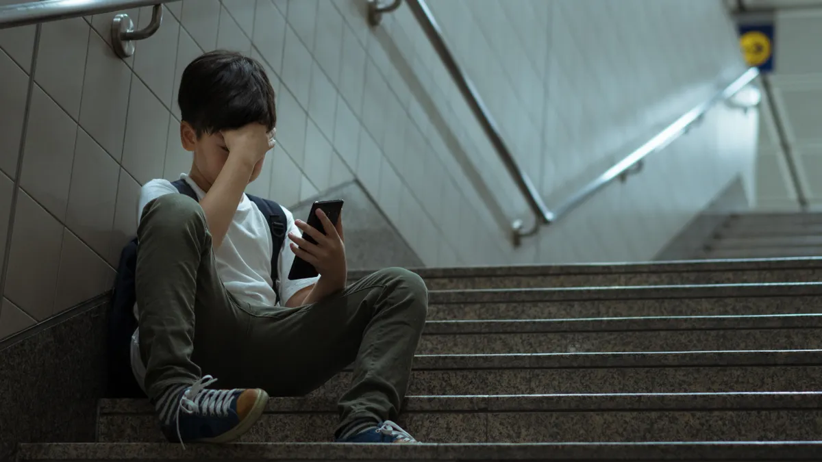 A young boy sits on a school staircase with a phone in his hand showing signs of stress