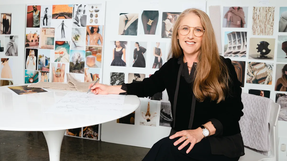 A person with long brown hair and glasses sits at a drawing table wearing a black top and pants and smiling at the camera