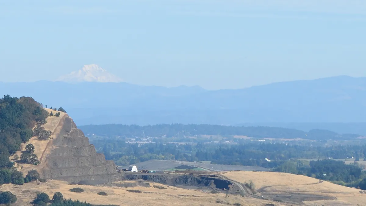 View of a stepped butte in foreground on left of image with landfill rising behind it and a snow-capped mountain in the background