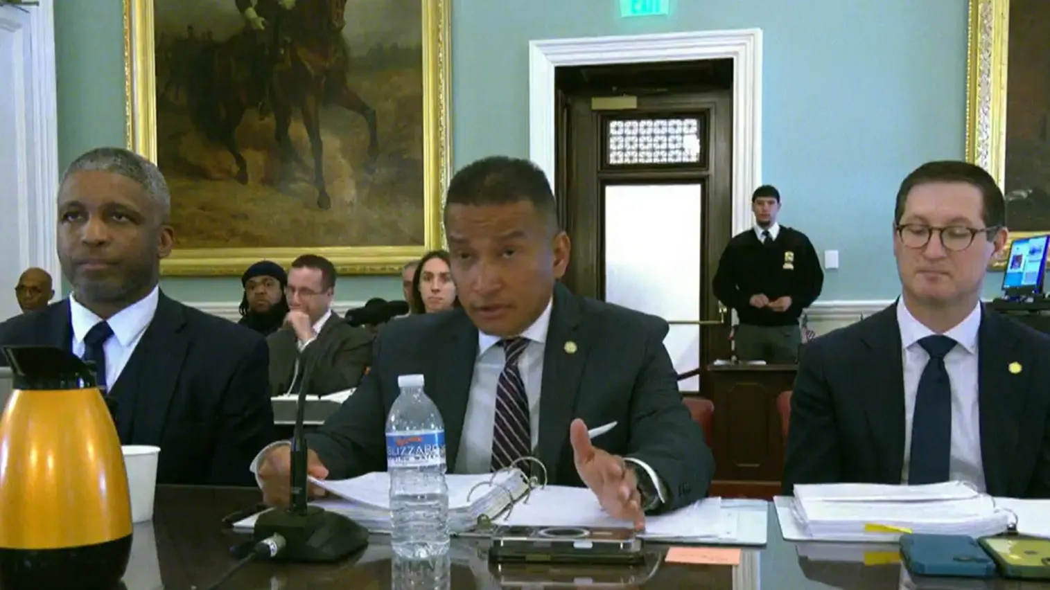 Three people sit at a table, with the one in the center talking into a microphone.