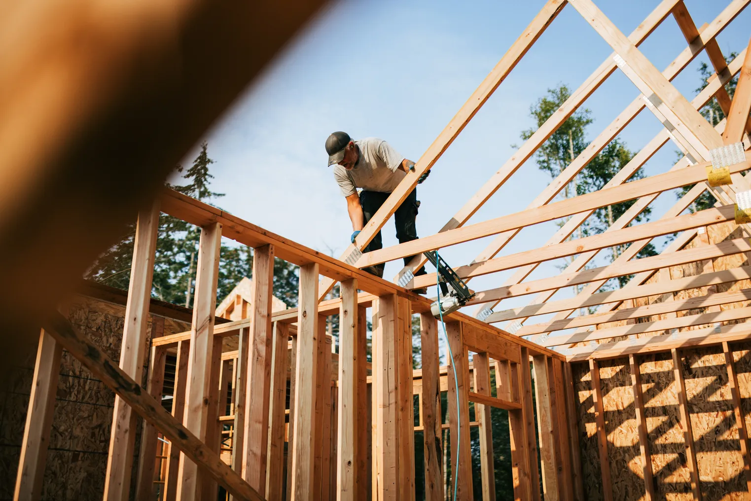 A person in a hat and gloves stands on the wooden beams of home under construction. The person holds a wooden beam.