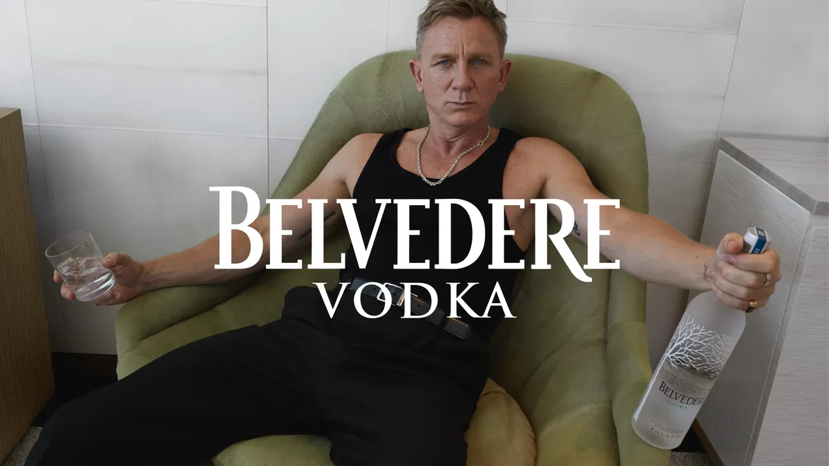 Actor Daniel Craig, wearing an all-black outfit, splays out on a green lounge chair while holding a bottle of Belvedere vodka in one hand and a glass tumbler in the other.