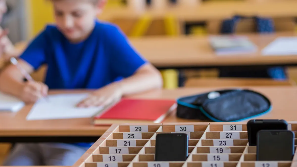 A box of cell phones sits in front of a student completing an assignment at their desk in a classroom.
