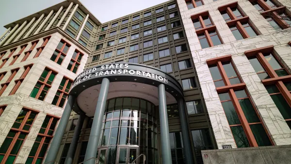 A large modern architectural design building with a grey stone façade and large glass windows displays the United States Department of Transportation sign above a cynical arch building entrance. 