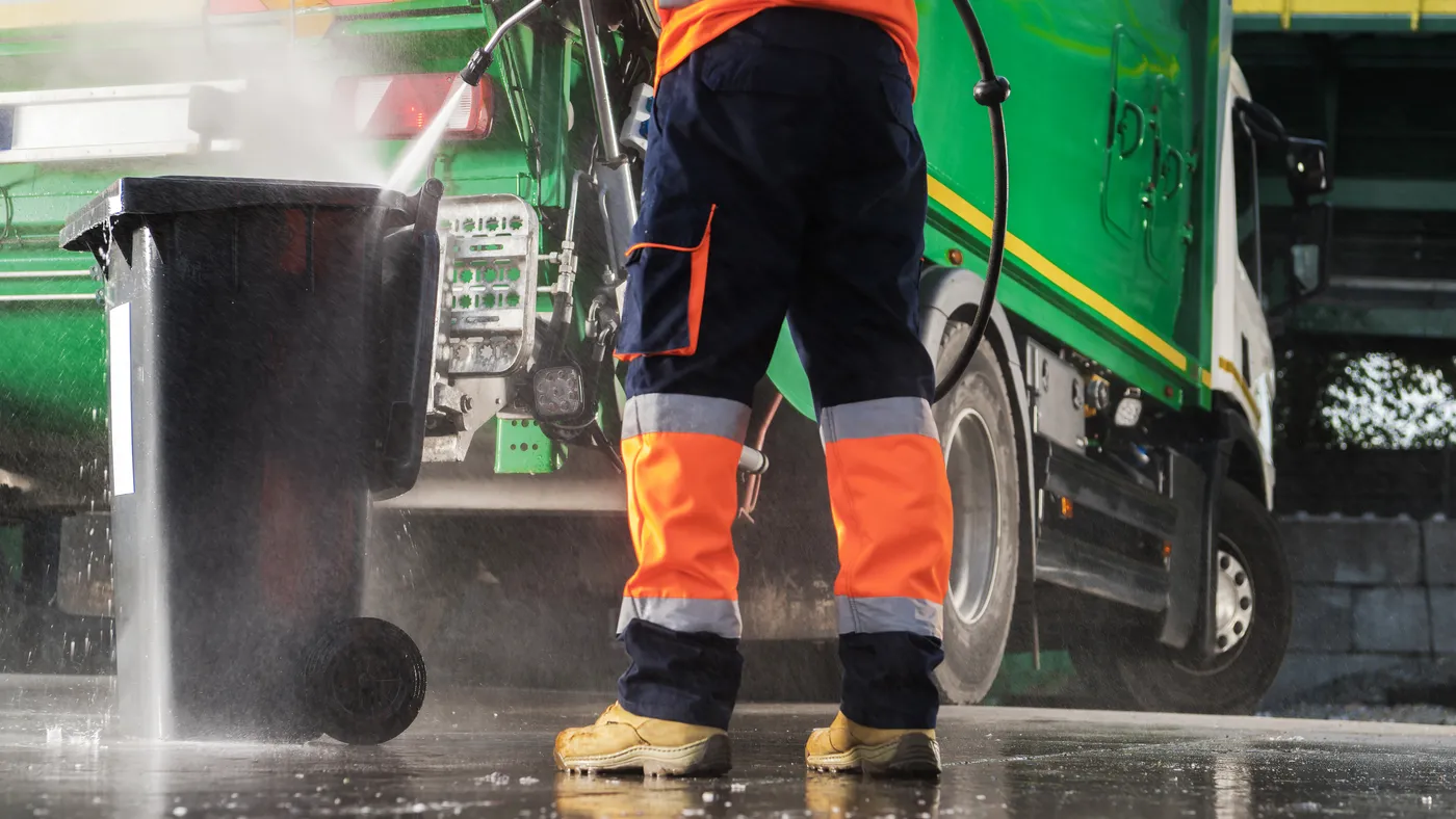 Person in work boots washing waste container next to green truck