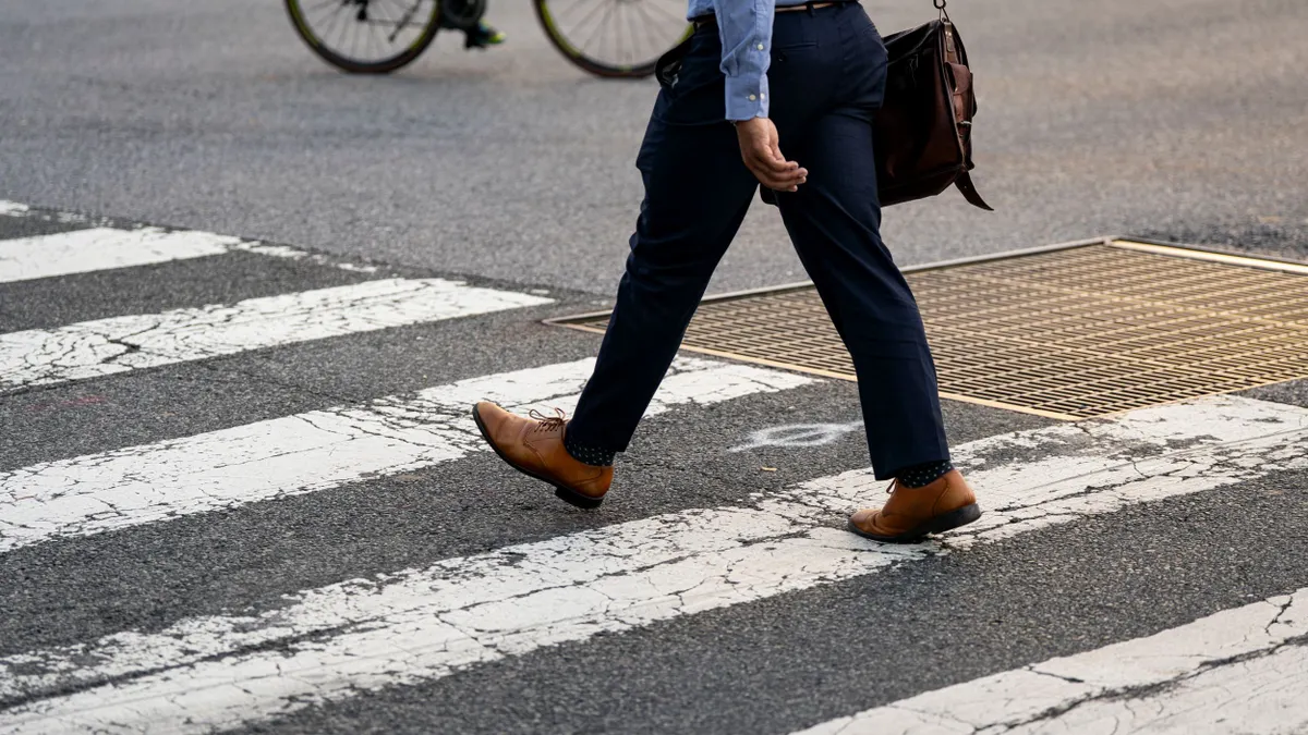 A person wearing slacks and dress shoes and carrying a shoulder bag moves through a crosswalk.