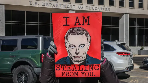 A person holds a sign outside of the U.S. Department of Education's building depicting Elon Musk's face and reading, in all capital letters, "I am stealing from you."