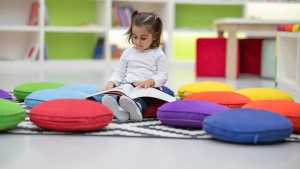 A young student is sitting in a room with books. The student is sitting among colorful cushions on the floor and looking at a book.